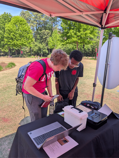 Two students play a mini excape room, standing outside at a table with boxes and a laptop.